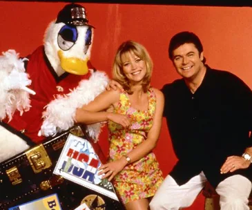 Three people, including someone in a duck costume, pose happily with a suitcase against an orange background.