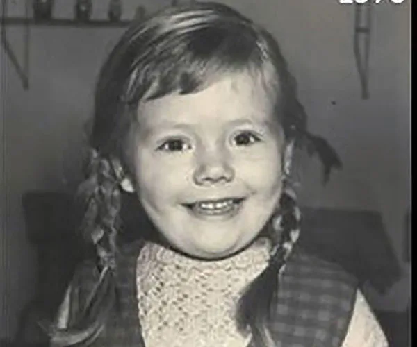 A young child with braided hair smiles at the camera in a black-and-white photo.