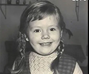 A young child with braided hair smiles at the camera in a black-and-white photo.