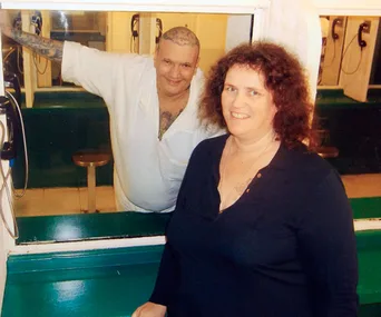In a prison visitation room, a man with tattoos behind glass smiles beside a woman in a dark shirt.