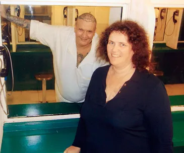 In a prison visitation room, a man with tattoos behind glass smiles beside a woman in a dark shirt.