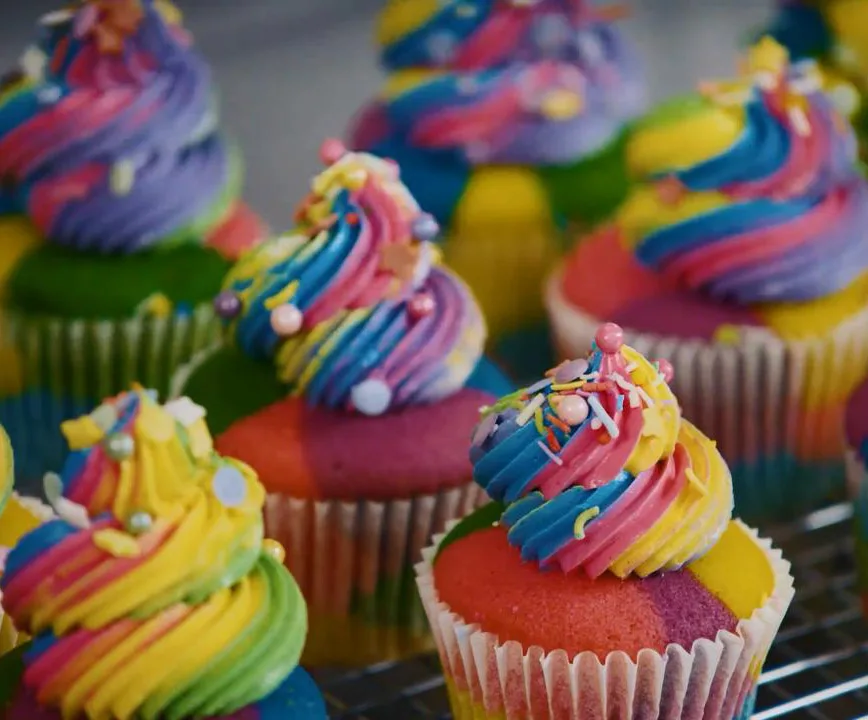 Colorful rainbow cupcakes with swirled frosting and sprinkles on top, arranged in rows.