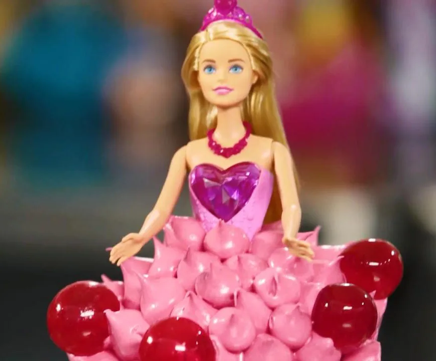Barbie doll cake with a pink dress made of frosting and candy, topped with a jewel-shaped candy and tiara.