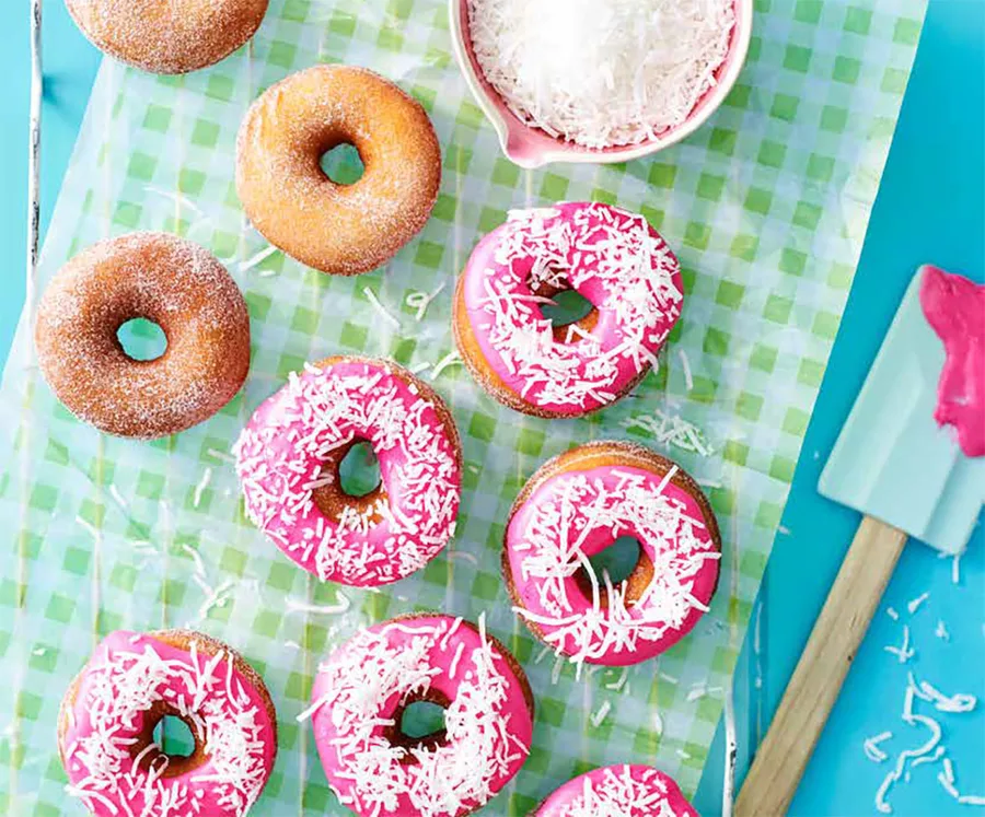 Pink-frosted doughnuts with coconut sprinkles on green gingham paper, next to a spatula and bowl of shredded coconut.