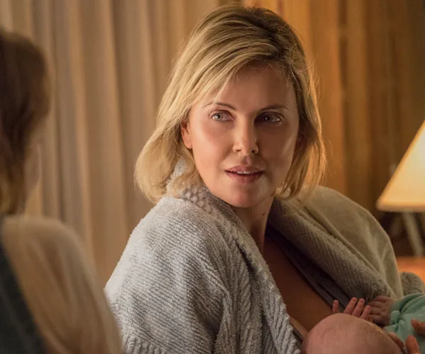 Charlize Theron's character, Marlo, from "Tully," sitting with a baby, wearing a cozy robe indoors.