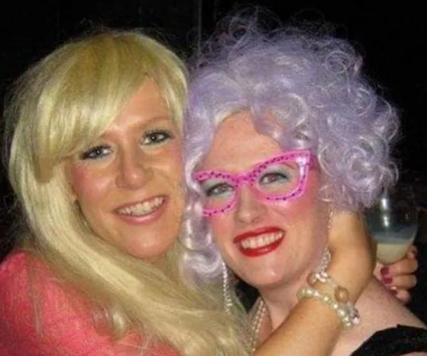 Two people smiling, one with blond hair, the other with curly lavender hair and pink glasses, embracing.