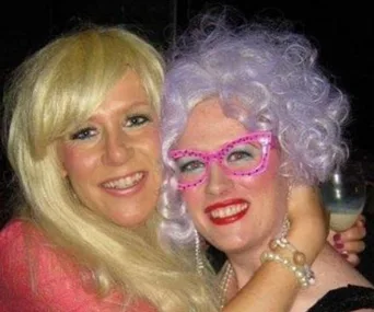 Two people smiling, one with blond hair, the other with curly lavender hair and pink glasses, embracing.