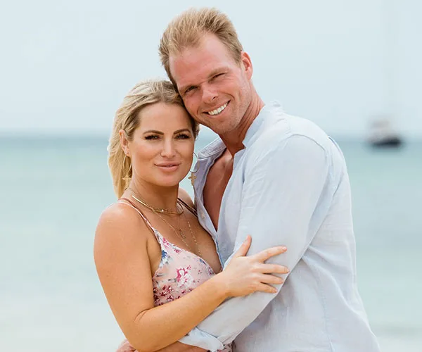 A couple embracing on a beach with the ocean in the background, both looking at the camera.