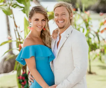 Couple smiling, outdoors; woman wears blue dress, man in light suit; tropical background.