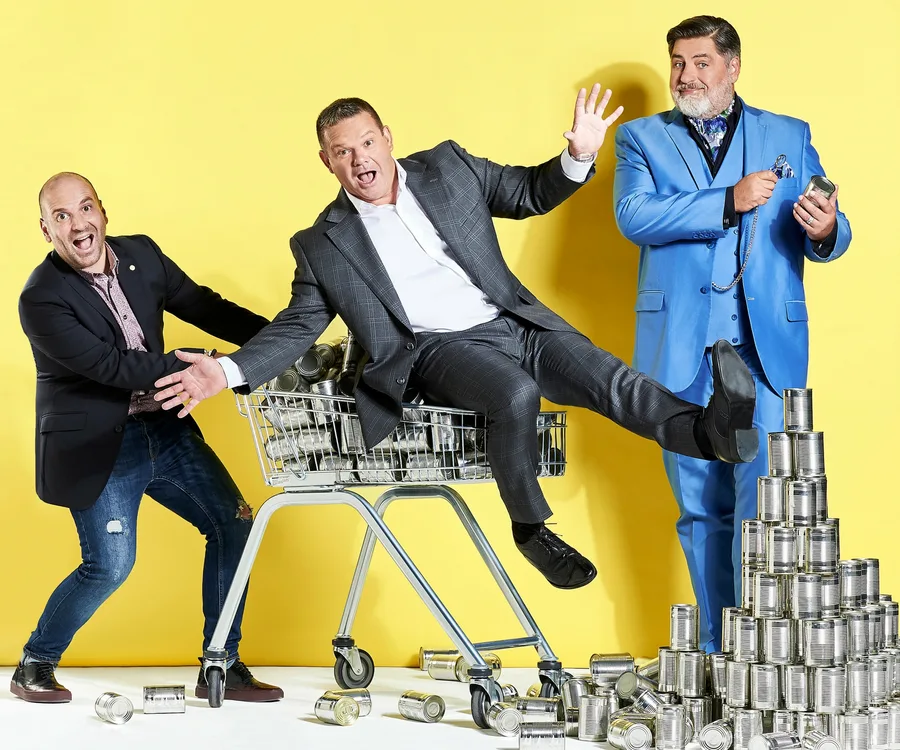 Three men with varied expressions pose with a shopping cart and stacked tin cans against a yellow background.