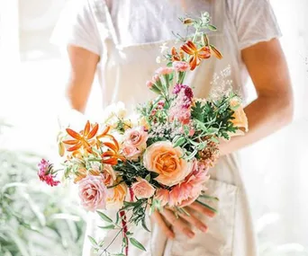 Bouquet of colorful flowers including roses and greenery held by a person in a white shirt and apron.