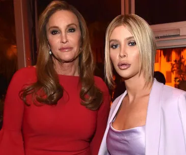 Two people standing together; one in a red dress, the other in a lilac outfit, at an indoor event.