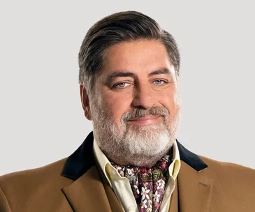 MasterChef judge in a brown coat and patterned scarf, smiling against a plain background.