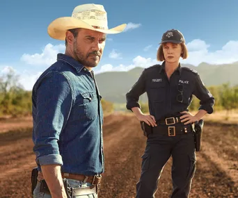 Two characters from "Mystery Road" stand on a dusty road; one in a blue shirt and cowboy hat, the other in a police uniform.