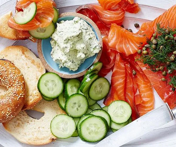 Bagel platter with smoked salmon, cucumber slices, herb cream cheese, and capers on a white tray.