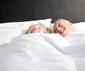 Elderly couple smiling under white bedsheets, lying side by side, showcasing happiness and companionship.