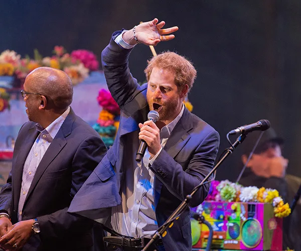 Prince Harry speaking on stage with a microphone, next to a man, colorful background.