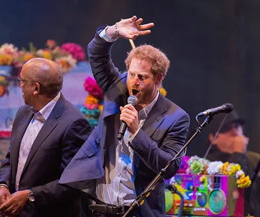 Prince Harry speaking on stage with a microphone, next to a man, colorful background.