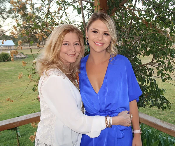 Two women smiling and embracing outdoors, one in a white outfit and the other in a blue dress, with greenery behind them.