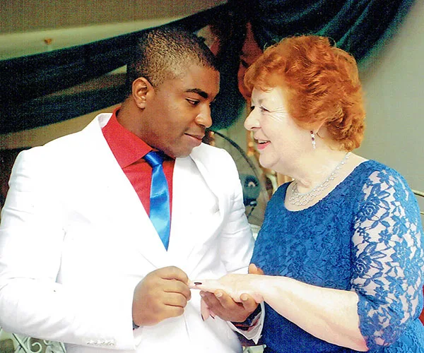 Young man and older woman in formal attire exchanging wedding rings, smiling warmly at each other.