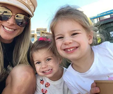 A woman wearing sunglasses smiles with two young girls, all posing happily outdoors.