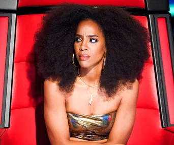 Judge on "The Voice" in a reflective dress, sitting in a red chair, with a thoughtful expression.