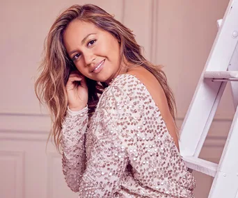 "Jessica Mauboy poses in a sparkling dress, smiling while seated beside a white ladder indoors."