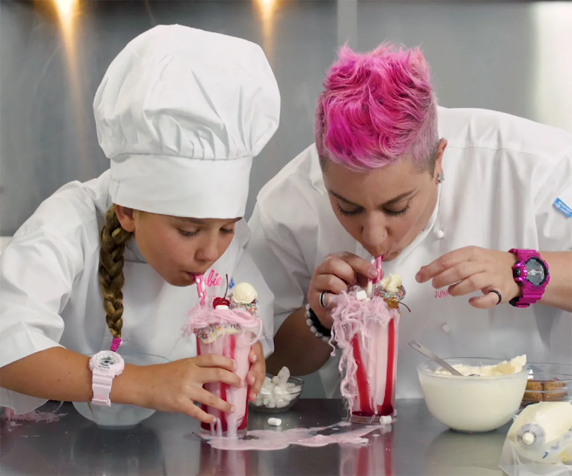 Chefs sipping extravagant pink milkshakes topped with cotton candy and ice cream in a kitchen setting.