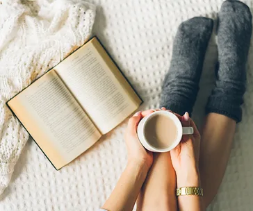 Person holding mug next to open book on cozy bed with grey socks.