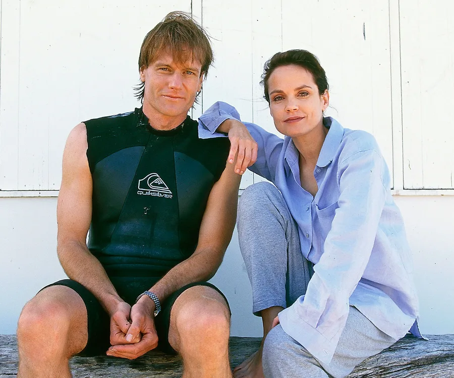 Two people sitting on a wooden bench, the man in a wetsuit and the woman in casual attire.
