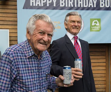 Two elderly men smiling, holding beer cans in front of a "Happy Birthday" banner.