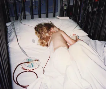 Young child lying in a hospital bed with medical equipment around, covered partially by a white sheet.