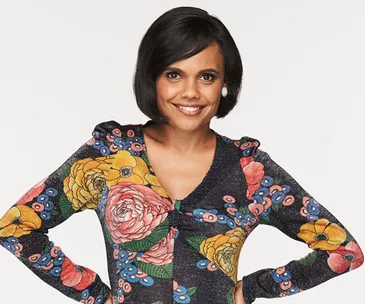 Miranda Tapsell smiling, wearing a colorful, floral-patterned dress against a plain background.