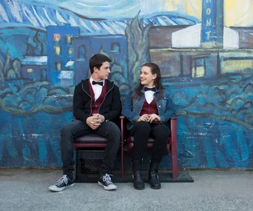 A young man and woman, dressed in formal attire, sit on chairs against a colorful mural background.