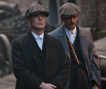 Two men in period attire, wearing caps and coats, stand looking forward; scene from "Peaky Blinders."