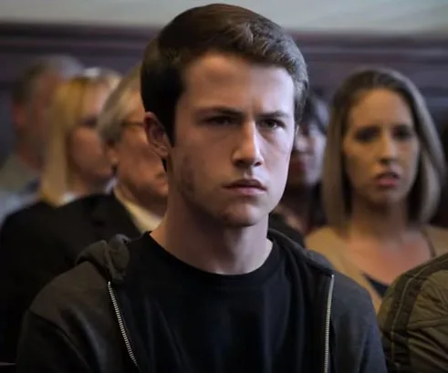 A young man with short hair and a serious expression sits in a courtroom, wearing a hoodie. People are seated in the background.