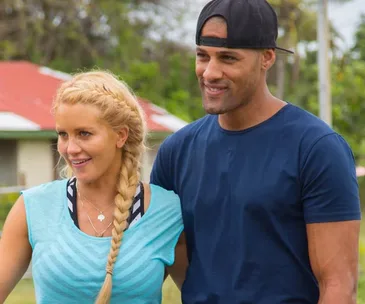 A smiling couple with the woman in a blue top and braided hair, and the man in a navy shirt and backwards cap.