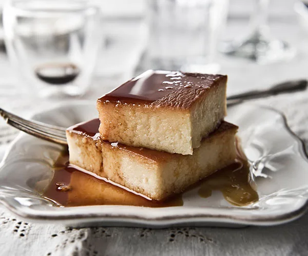 Two square slices of flan on a plate with caramel sauce, fork beside it.