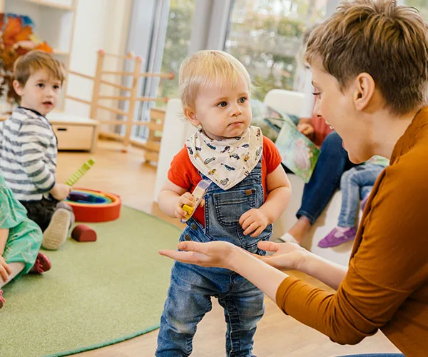 Toddler in overalls stands with caregiver in a playful, colorful childcare setting.