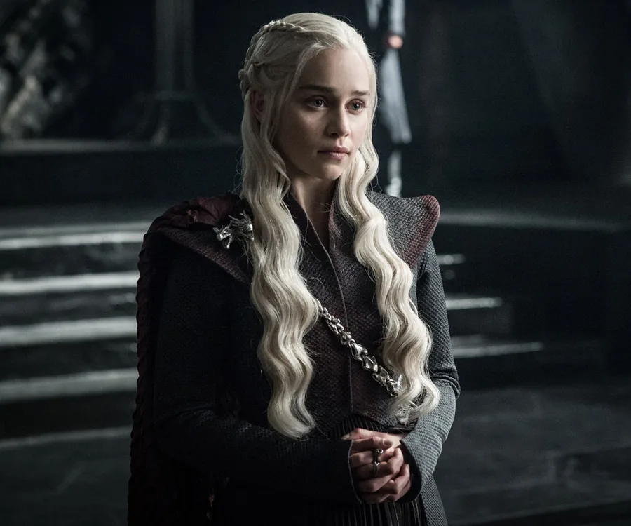 "Daenerys Targaryen with long braid in armor-like outfit, standing in a dimly lit room from Game of Thrones, Season 8."