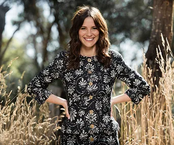 Young woman in patterned dress smiles while standing in a field with tall grass and trees in the background.
