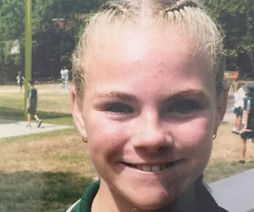 Young girl with blonde braided hair smiling outdoors at a park, other children visible in the background.