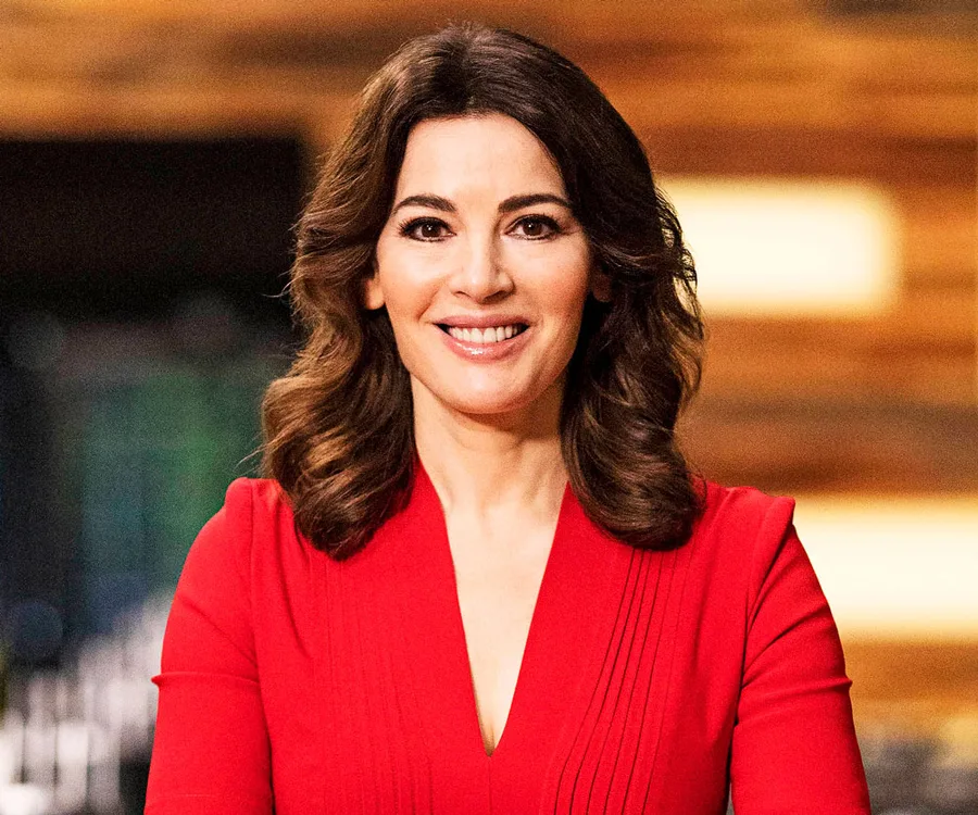 A woman with long dark hair wearing a red dress smiles warmly against a blurred indoor background.