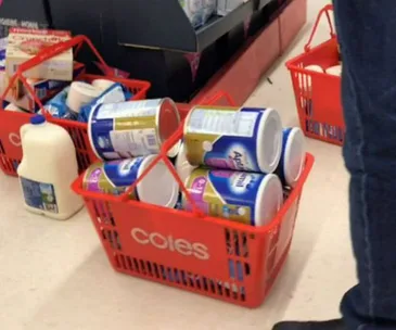 Red Coles shopping basket filled with Aptamil baby formula cans on a store floor.