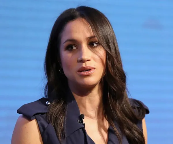 A woman with long dark hair and a serious expression, wearing a dark sleeveless top, against a blue backdrop.