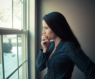 Woman with long hair looking out a window, deep in thought, with snow visible outside.