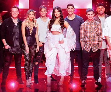 Group of contestants from "The Voice Australia" Top 12 Wildcards smiling on stage under bright lights.