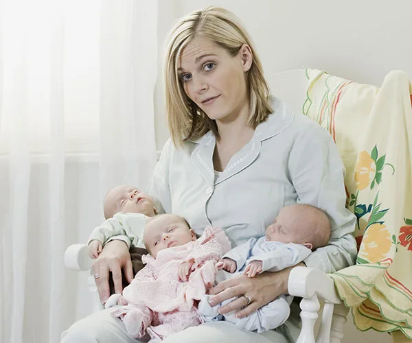 A woman sits in a chair holding three sleeping babies, looking slightly overwhelmed but affectionate.