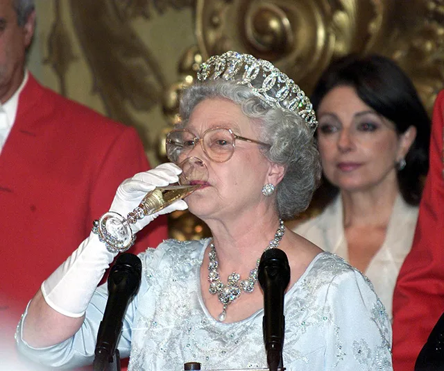 An elderly woman in a tiara and gloves drinking champagne at a formal event.