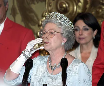 An elderly woman in a tiara and gloves drinking champagne at a formal event.